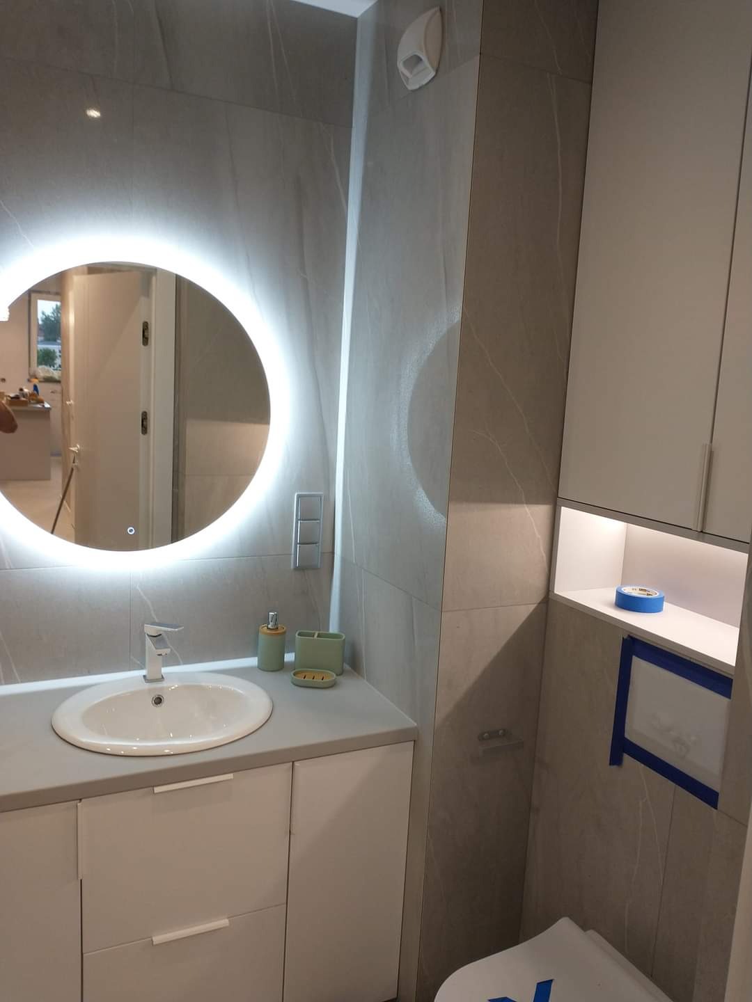 Nowoczesna łazienka z podświetlanym lustrem, umywalką na szafce, szarą glazurą imitującą marmur i zabudowaną toaletą z niebieską taśmą zabezpieczającą.