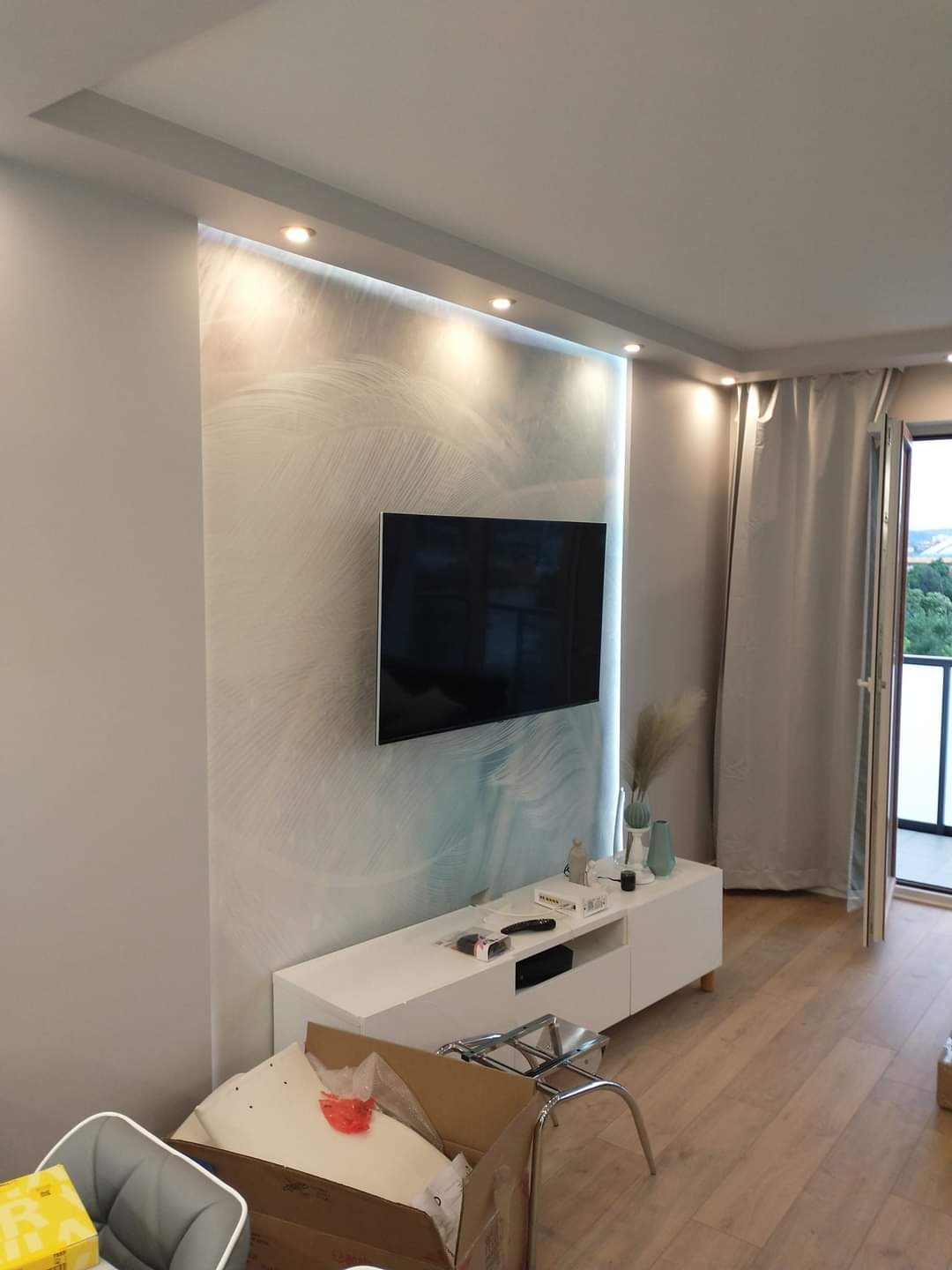 Salon po remoncie z telewizorem na ścianie z dekoracyjną tapetą, oświetleniem punktowym i jasną podłogą z paneli. Częściowo rozpakowane meble w kartonach na pierwszym planie.