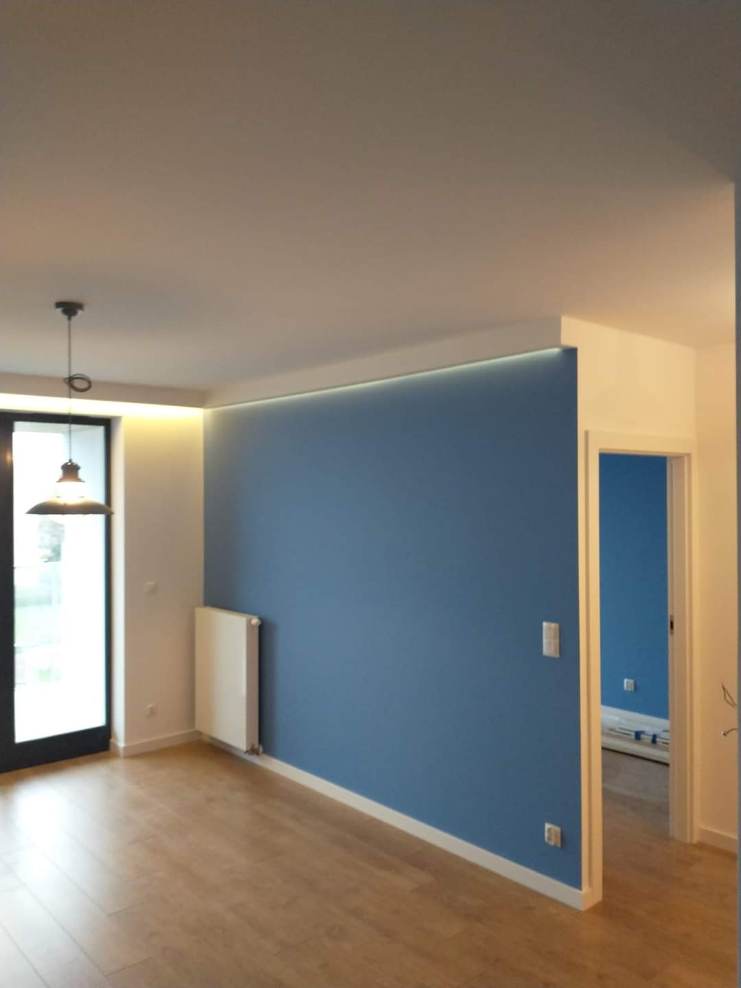 Pokój z pomalowaną na niebiesko ścianą, oświetleniem LED podsufitowym, drewnianą podłogą i czarnymi drzwiami balkonowymi. W tle widoczny fragment innego, również niebieskiego pokoju.