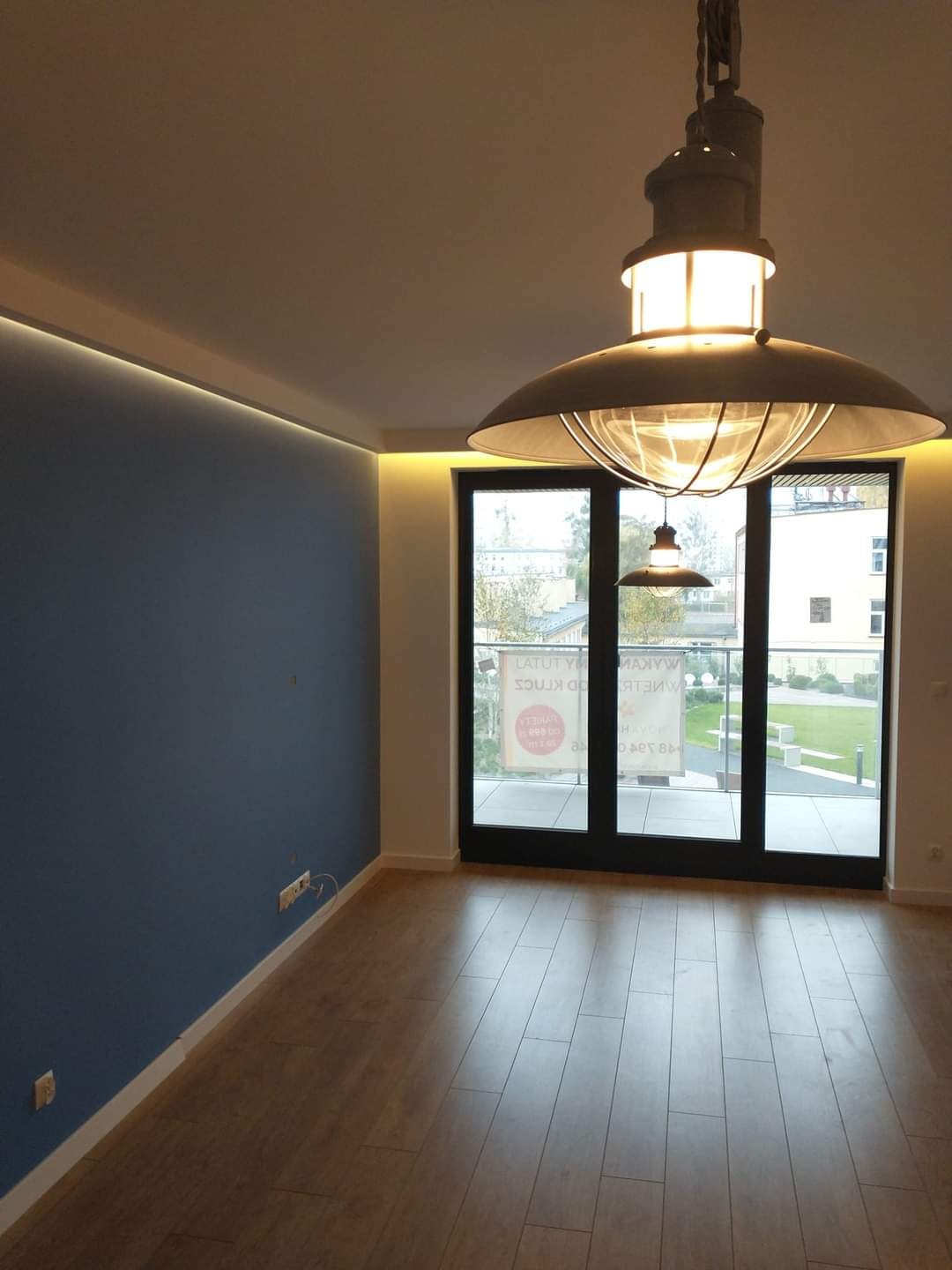 Pusta przestrzeń po remoncie: ściana pomalowana na niebiesko z listwą LED pod sufitem, parkiet ułożony na podłodze, duże okno balkonowe i designerska lampa w stylu industrialnym.