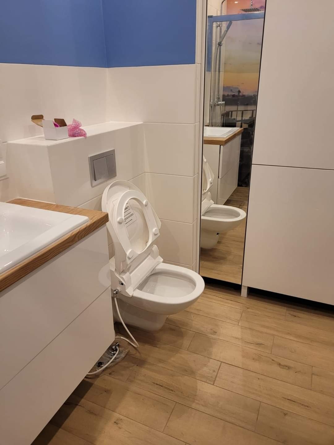 Nowoczesna toaleta z deską bidetową, biała armatura łazienkowa, drewnopodobne płytki na podłodze i niebieski akcent kolorystyczny na ścianie.