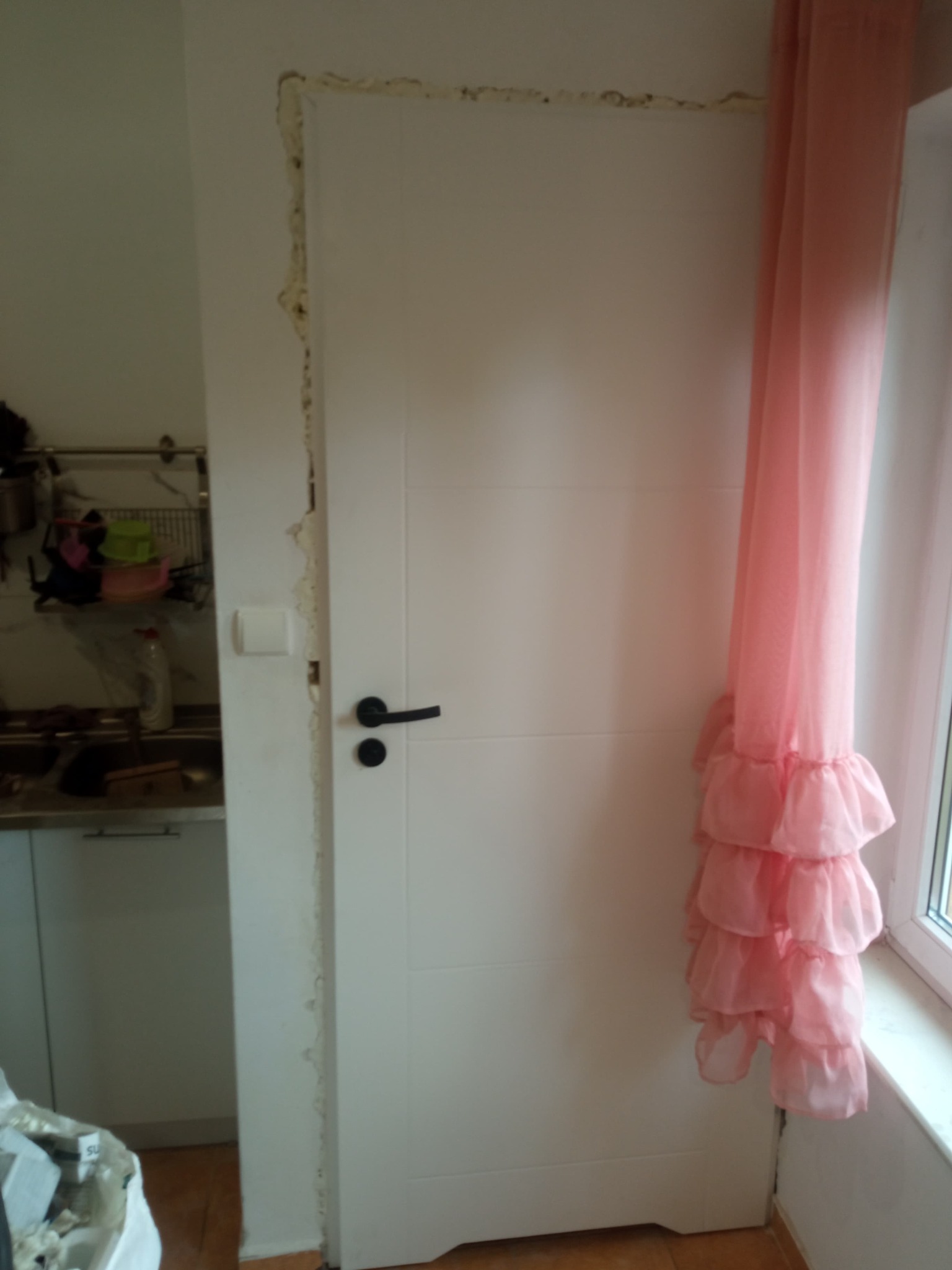 Białe drzwi wewnętrzne z czarną klamką, osadzone w ościeżnicy z widocznymi śladami pianki montażowej i szpachli, obok okna z różową firanką w falbany.