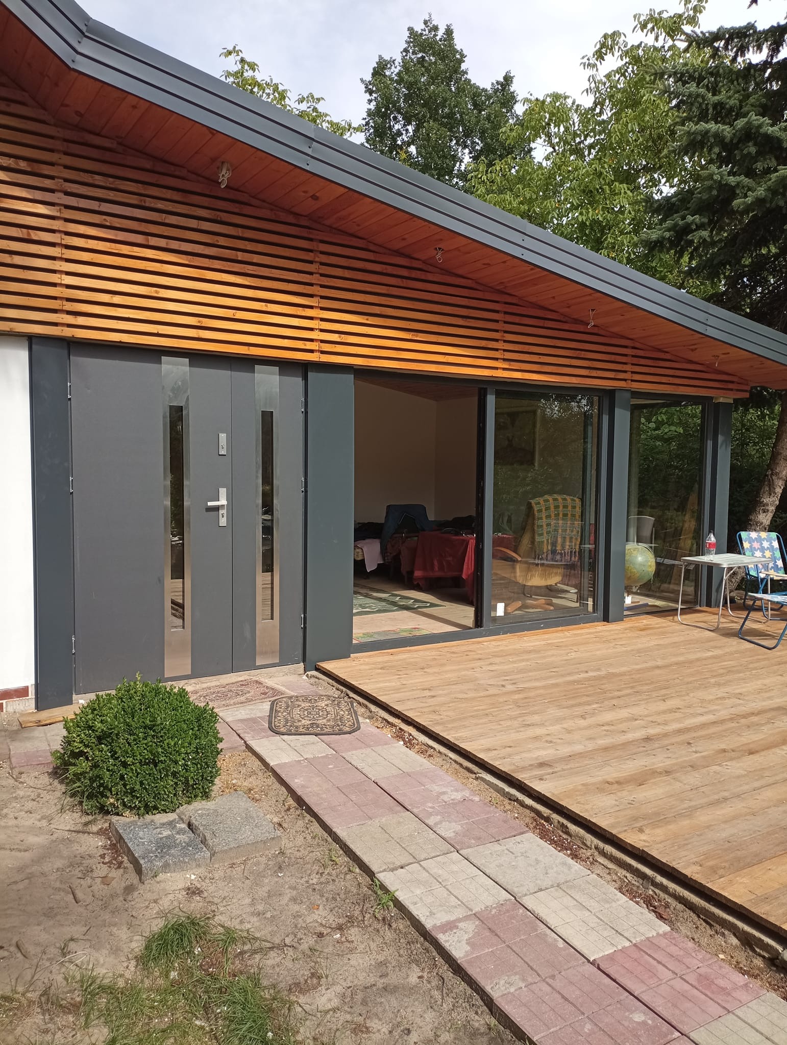 Nowoczesny budynek z drewnianą elewacją i tarasem, szare drzwi przesuwne z przeszkleniami, widok na wnętrze z meblami i dekoracjami.