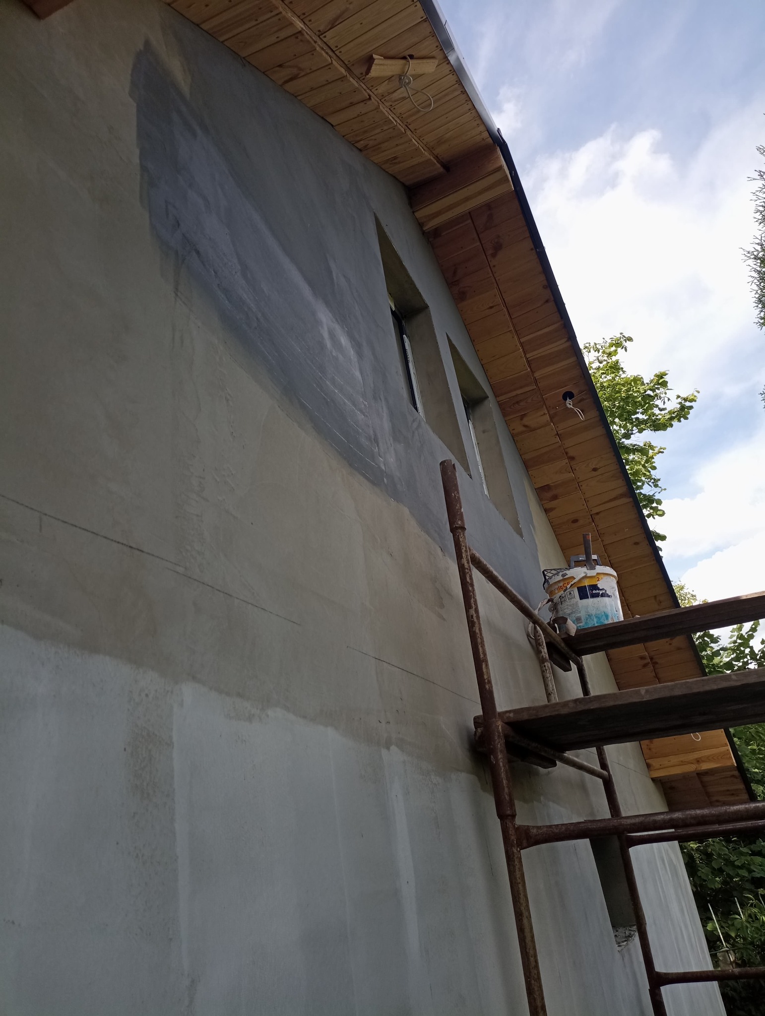Ściana budynku w trakcie przygotowania do malowania, widoczne ślady szpachlowania i gruntowania, rusztowanie z wiadrem farby obok okien.