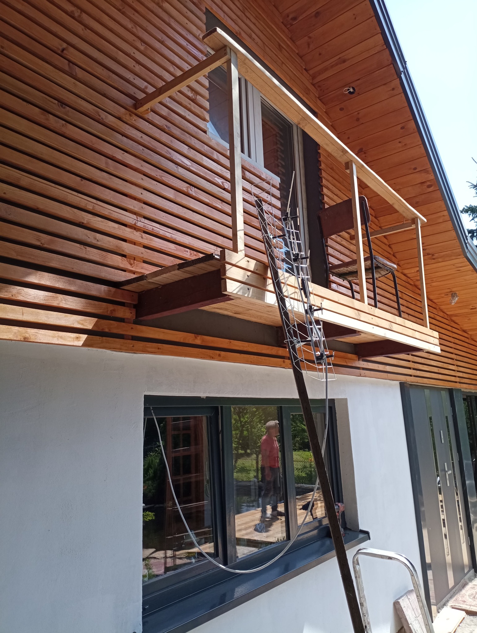 Dom z nową drewnianą elewacją i dobudowanym balkonem, widoczna antena TV oparta o ścianę, odbicie osoby w oknie.