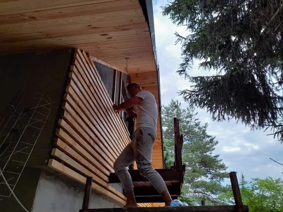 Pracownik na rusztowaniu mocuje drewniane listwy elewacyjne do ściany budynku, widoczne elementy konstrukcyjne domu i otaczająca zieleń.