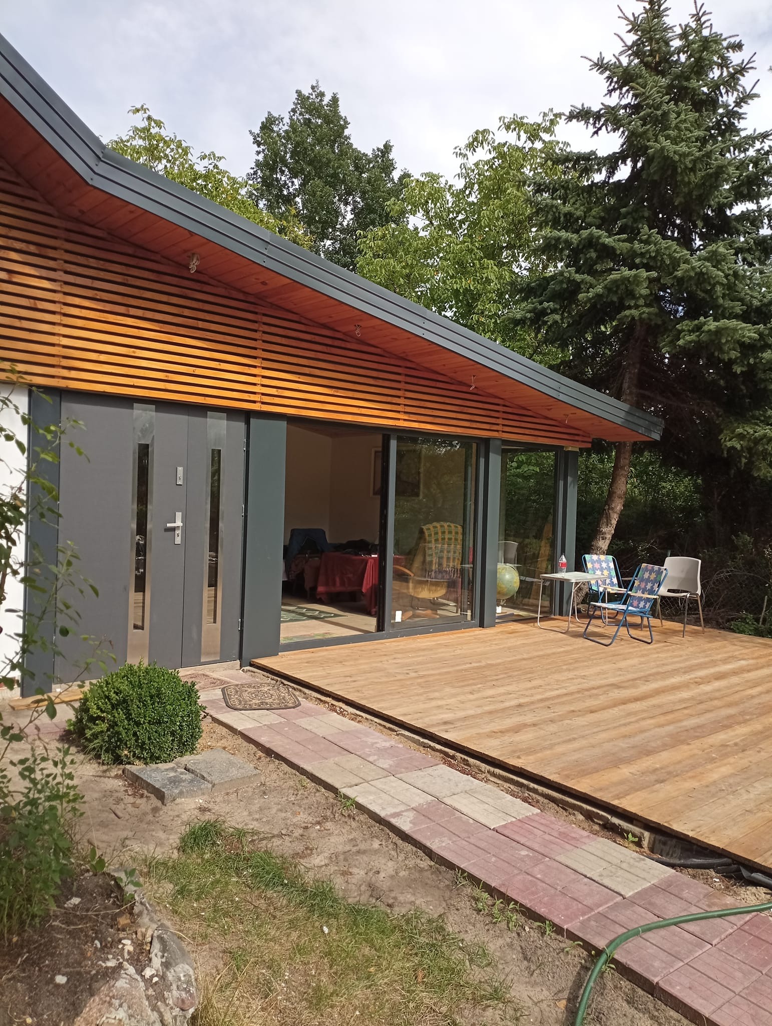 Nowoczesny parterowy dom z drewnianą elewacją i dużymi oknami, taras z desek, meble ogrodowe na zewnątrz.