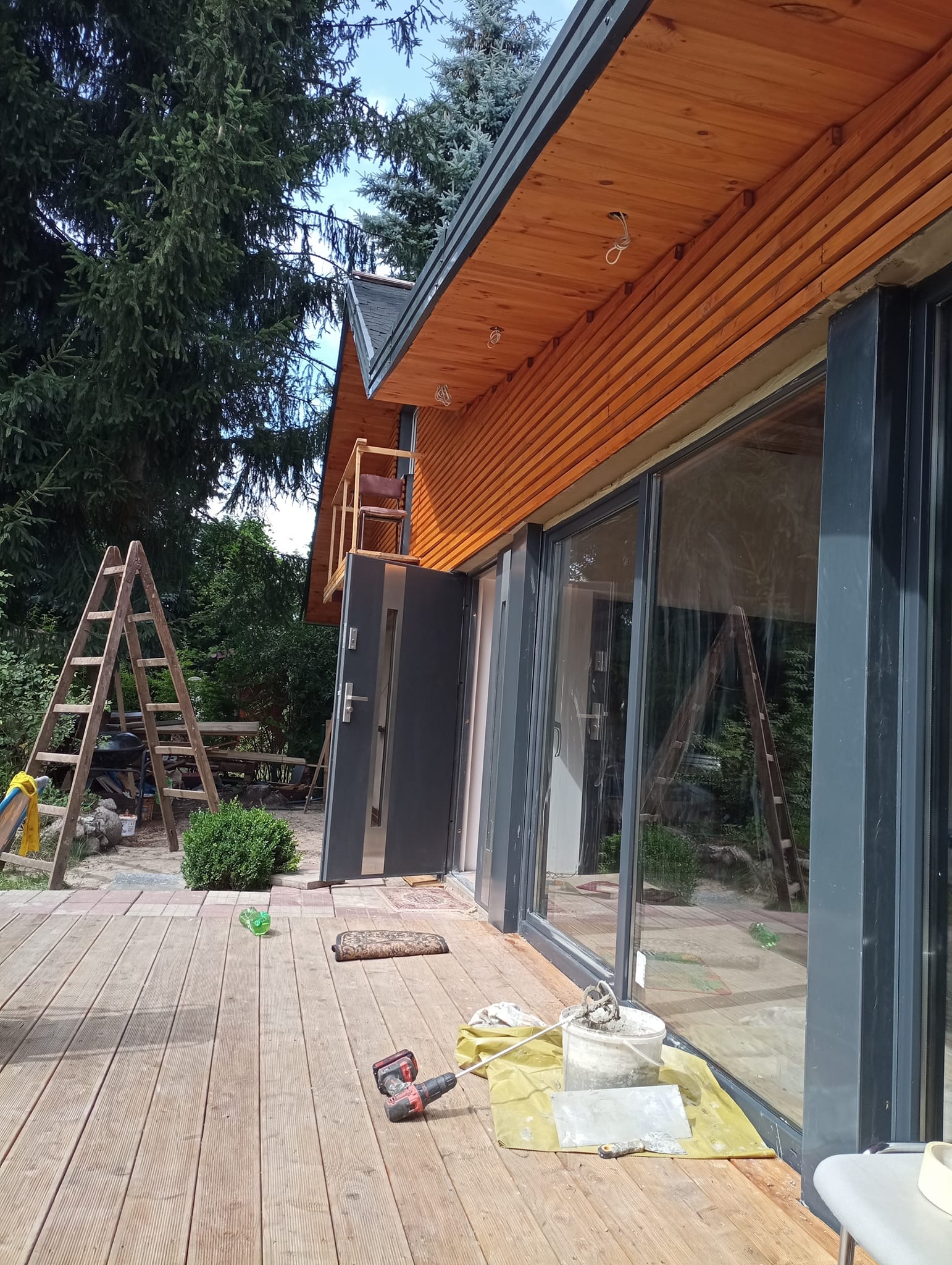 Dom z drewnianą elewacją w trakcie wykańczania, widoczne narzędzia, drabina i wiadro z zaprawą na tarasie.