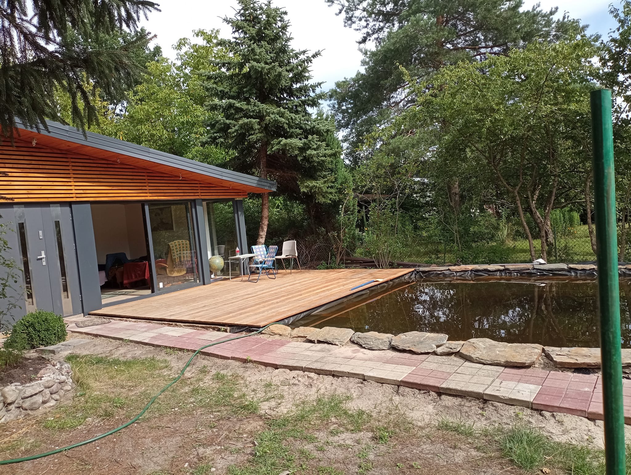 Nowoczesny domek letniskowy z drewnianym tarasem nad oczkiem wodnym, otoczony zielenią i brukowaną ścieżką