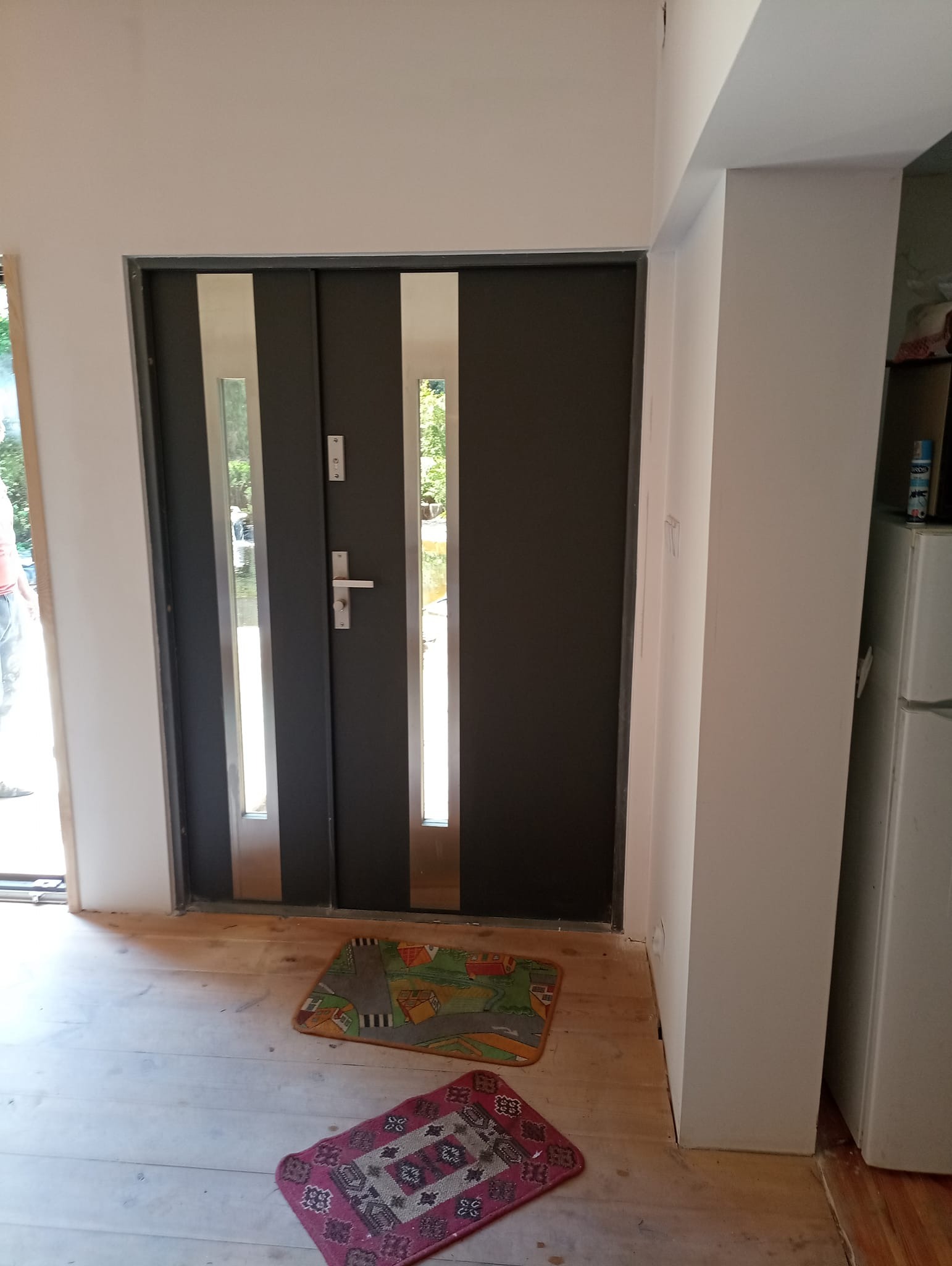 Nowe, grafitowe drzwi wejściowe z przeszkleniami, zamontowane w surowym stanie wykończeniowym, widoczne drewniane podłoże i dwa dywaniki.
