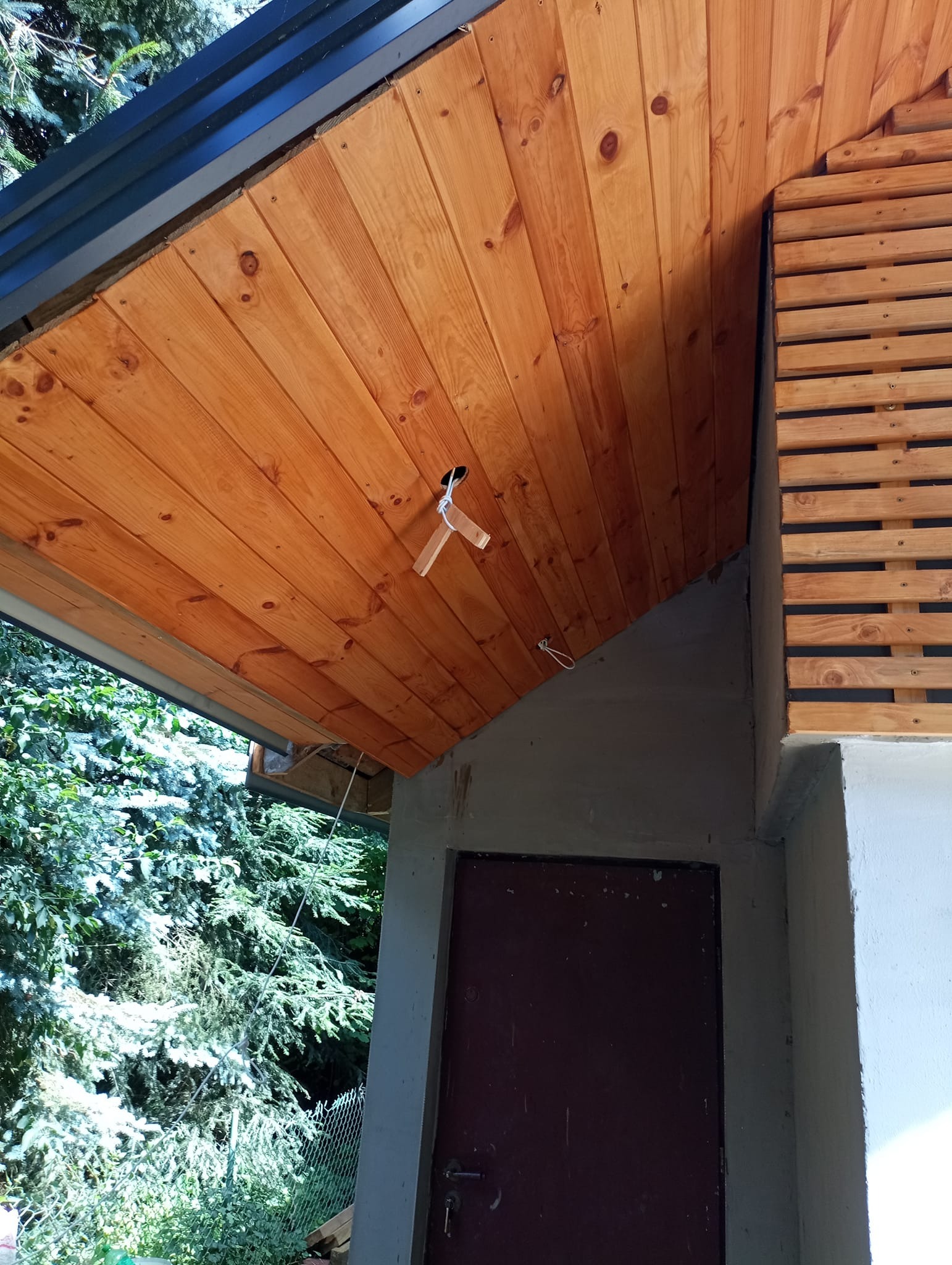 Drewniana podbitka dachowa z widocznym otworem na oświetlenie, drewniana elewacja oraz wejście do budynku w stanie surowym.