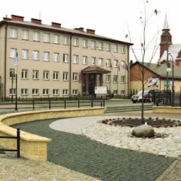 Kwadratowa nawierzchnia z ciemnej kostki brukowej przechodząca w okrąg z jasnych kamieni otaczający drzewo z kamieniem u podstawy, z budynkiem w tle.