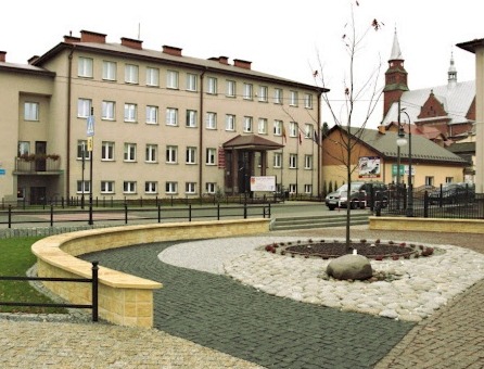 Kwadratowa nawierzchnia z ciemnej kostki brukowej przechodząca w okrąg z jasnych kamieni otaczający drzewo z kamieniem u podstawy, z budynkiem w tle.