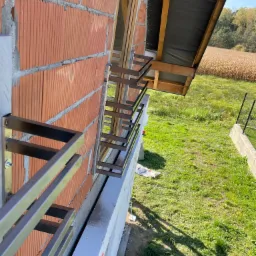 Montaż stalowej balustrady okiennej z profili zamkniętych, widok z zewnątrz na surową ścianę z cegieł ceramicznych i ocieplenie styropianem, w tle pole kukurydzy.