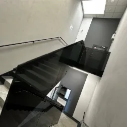 Schody wykończone płytkami imitującymi marmur, zabezpieczone szklaną balustradą z czarnymi detalami i czarną poręczą mocowaną do ściany, widok z góry.