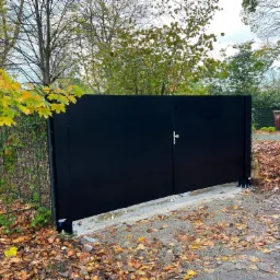 Nowoczesna, dwuskrzydłowa brama wjazdowa, pomalowana na czarno, z minimalistyczną klamką, zamontowana na betonowej podstawie, otoczona przez opadłe liście i jesienną roślinność.