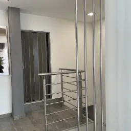 Nowoczesna klatka schodowa z metalową balustradą o minimalistycznym designie, z pionowymi, cienkimi słupkami i poziomymi rurkami, na tle białych ścian i szarych płytek podłogowych.