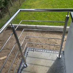 Błyszcząca, metalowa balustrada z poziomymi prętami, zamontowana na betonowym tarasie w trakcie budowy, widok z góry na surowy teren i zielony trawnik w tle.