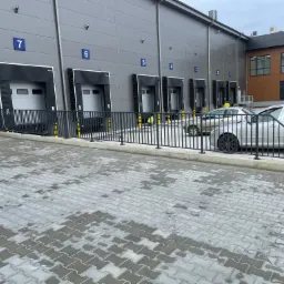Nowoczesna hala magazynowa z rampami załadunkowymi oznaczonymi numerami, zabezpieczona czarną, metalową barierką, z dwoma zaparkowanymi samochodami osobowymi.