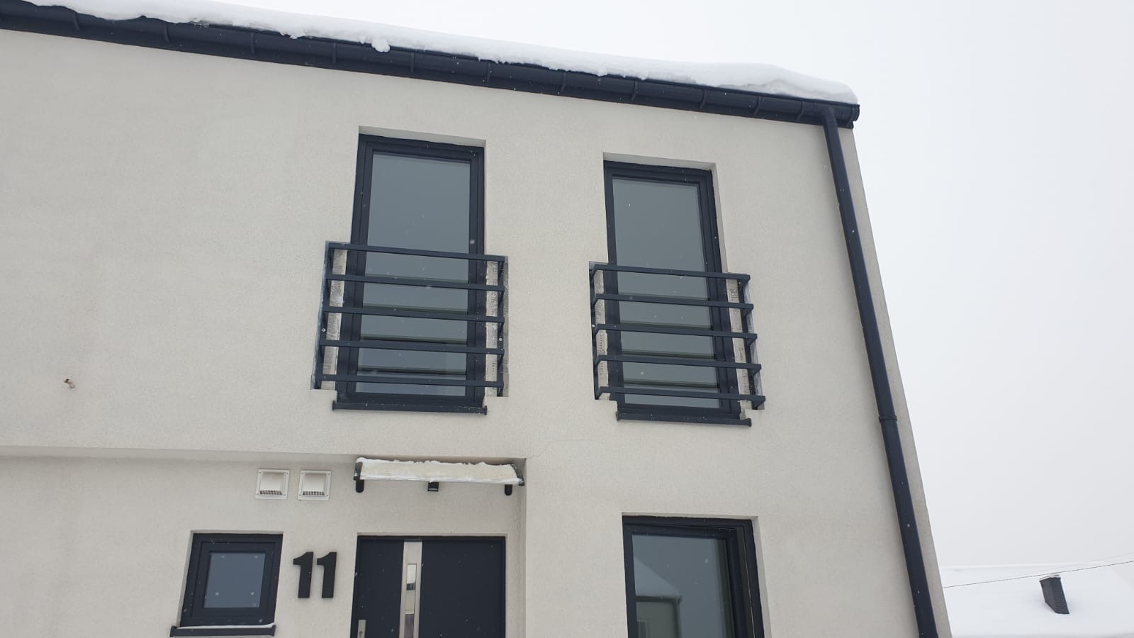 Elewacja dwupiętrowego domu z numerem 11 nad wejściem, okna z czarnymi ramami zabezpieczone poziomymi, czarnymi barierami, pokryte śniegiem.
