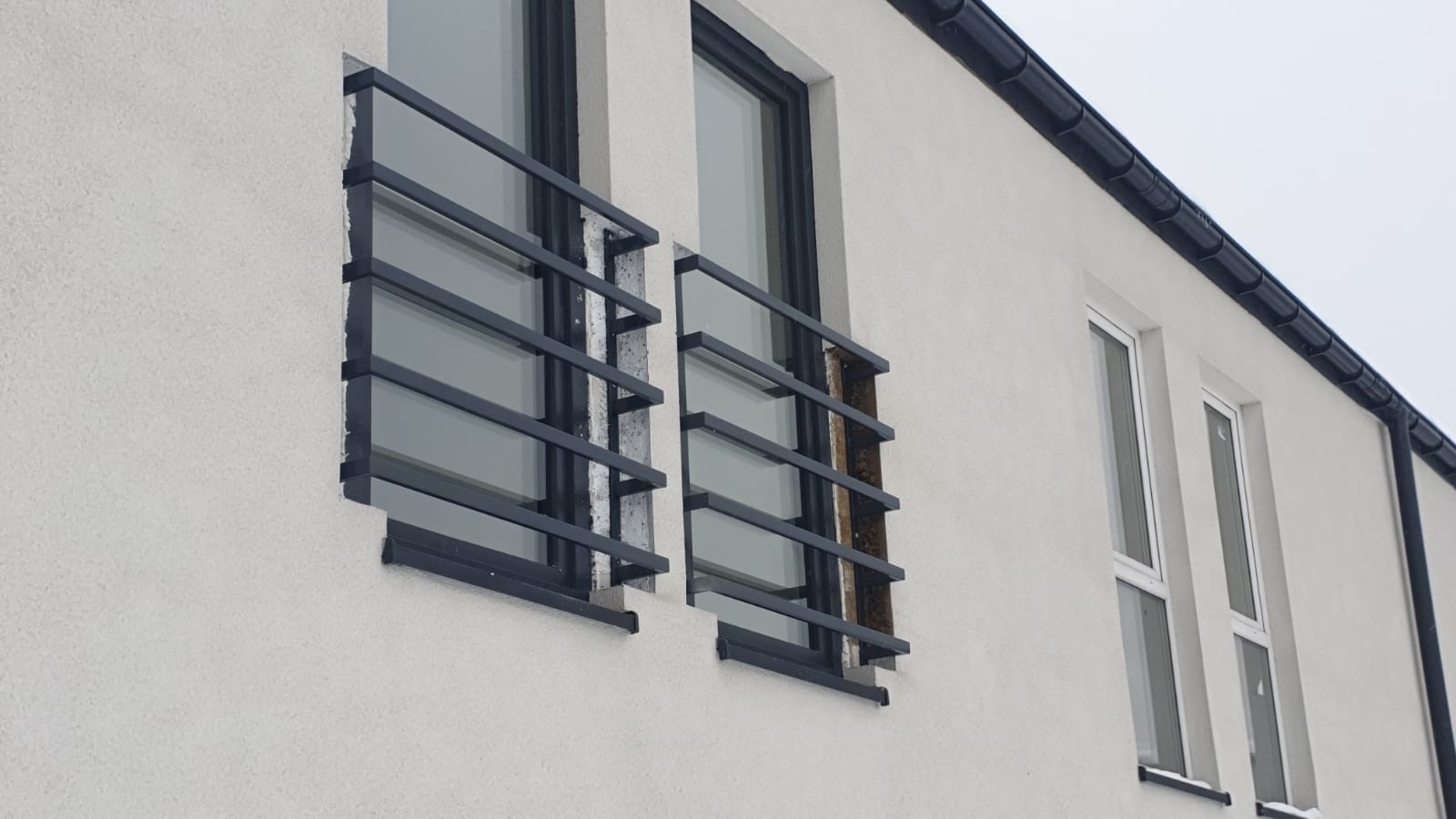 Elewacja budynku z dwoma oknami zabezpieczonymi poziomymi, czarnymi balustradami stalowymi; widoczne fragmenty uszkodzonego ocieplenia przy mocowaniu balustrad.