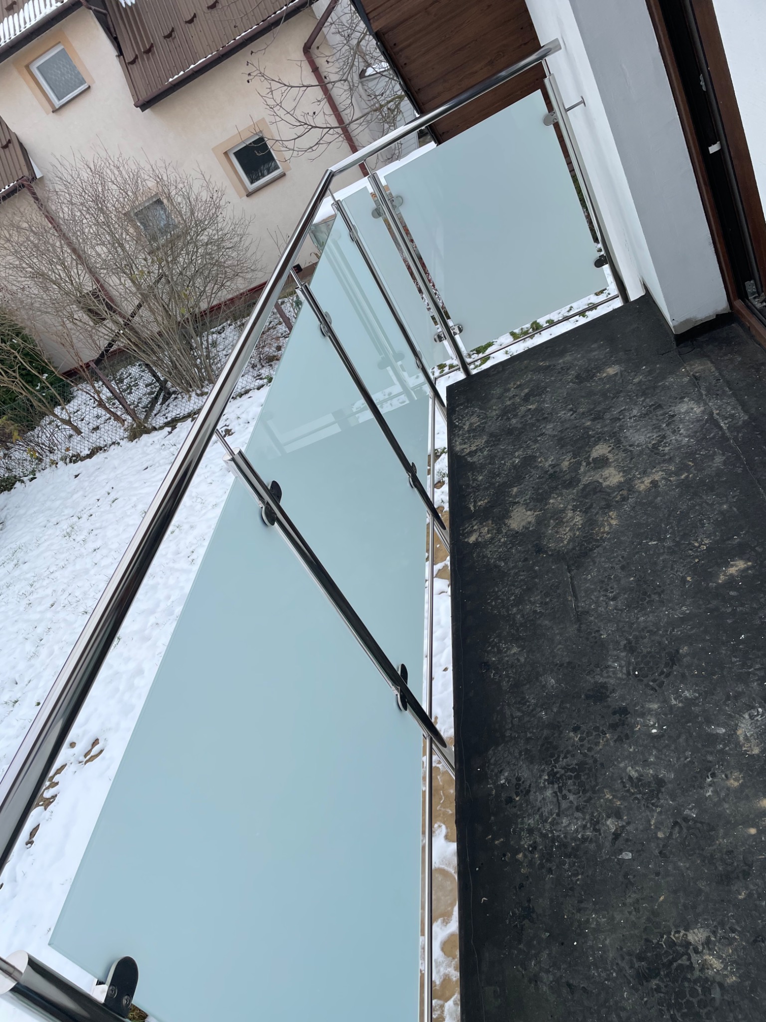 Zewnętrzna balustrada ze szkła mlecznego i stali nierdzewnej przy wejściu do domu, widok z góry, zimowy krajobraz z resztkami śniegu w tle.