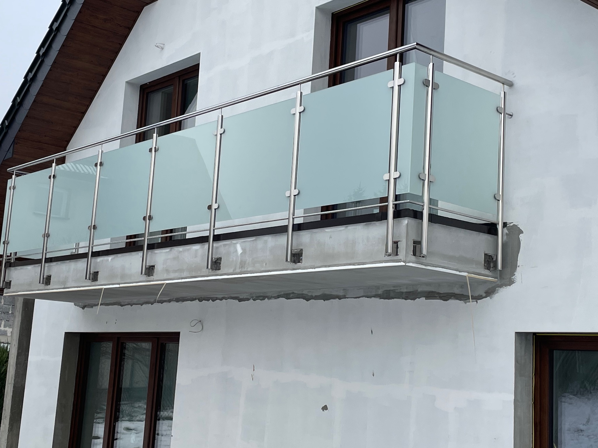 Balkon z balustradą ze szkła mlecznego i stali nierdzewnej, widoczne niedokończone prace wykończeniowe na elewacji budynku.