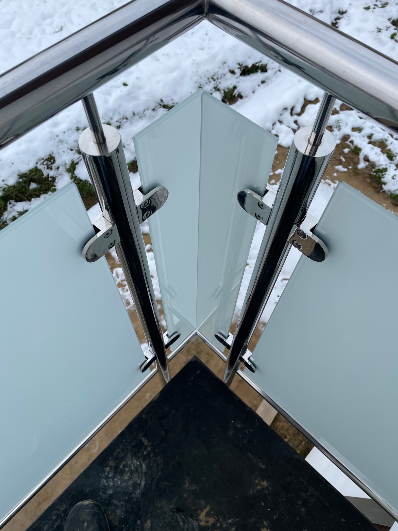 Widok z góry na narożnik balkonu z mlecznymi szklanymi panelami i chromowanymi słupkami, na tle ośnieżonego ogrodu.