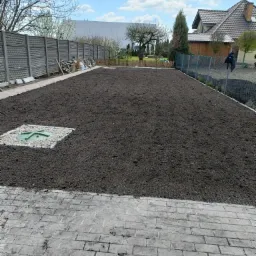 Przygotowany teren pod trawnik z widocznym włazem studzienki kanalizacyjnej otoczonym białym grysem, obrzeżony kostką brukową i ogrodzeniem z siatki oraz betonowych płyt.