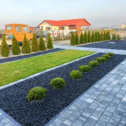 Nowoczesny ogród z geometrycznym układem: szara kostka brukowa, czarny grys, trawnik i szpaler tuj, z żółtym samochodem dostawczym w tle.