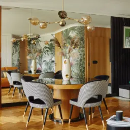 Jadalnia w stylu eklektycznym z okrągłym stołem, krzesłami w pepitkę, parkietem w jodełkę, lustrem na ścianie i lampą sufitową z kulistymi kloszami. W tle tapeta z motywem roślinnym i drewniana...