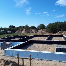 PITBUDD BUILDING CONTRACTORS - Przygotowany zbrojony fundament pod budowę domu jednorodzinnego z izolacją termiczną, widoczne bloczki betonowe i drewniane podpory, w tle usypana ziemia i zieleń.