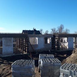 PITBUDD BUILDING CONTRACTORS - Wznoszenie ścian parteru domu z betonu komórkowego Ytong, widoczne zbrojenie nadproży, palety bloczków na pierwszym planie, pracownik na górze budynku macha ręką.