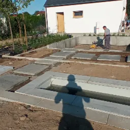 Prace ziemne i betonowe przy budowie ogrodu z elementami wodnymi; pracownik zagęszcza podłoże ubijarką wibracyjną, widoczne betonowe koryta i płyty chodnikowe.