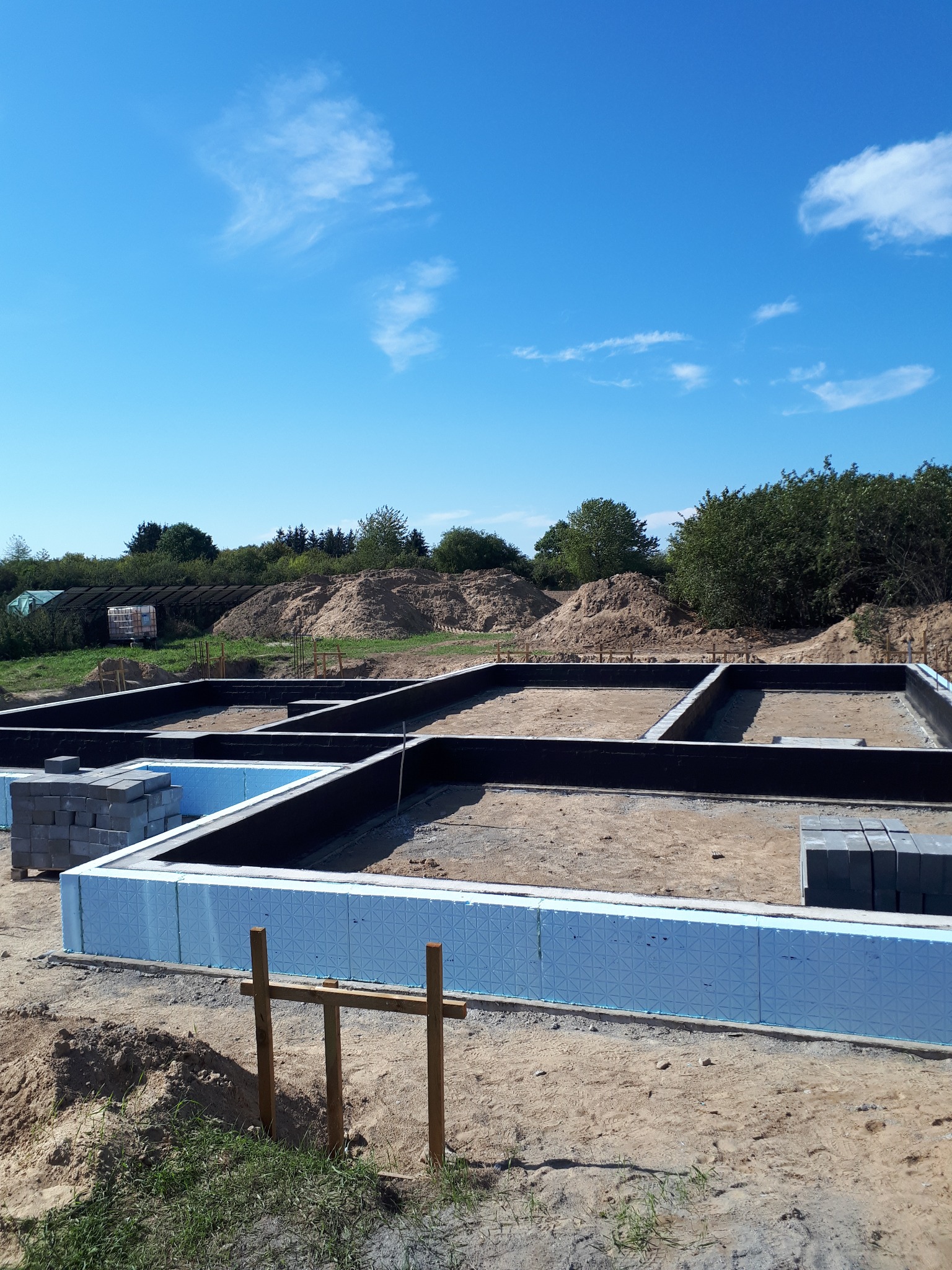 Przygotowany zbrojony fundament pod budowę domu jednorodzinnego z izolacją termiczną, widoczne bloczki betonowe i drewniane podpory, w tle usypana ziemia i zieleń.