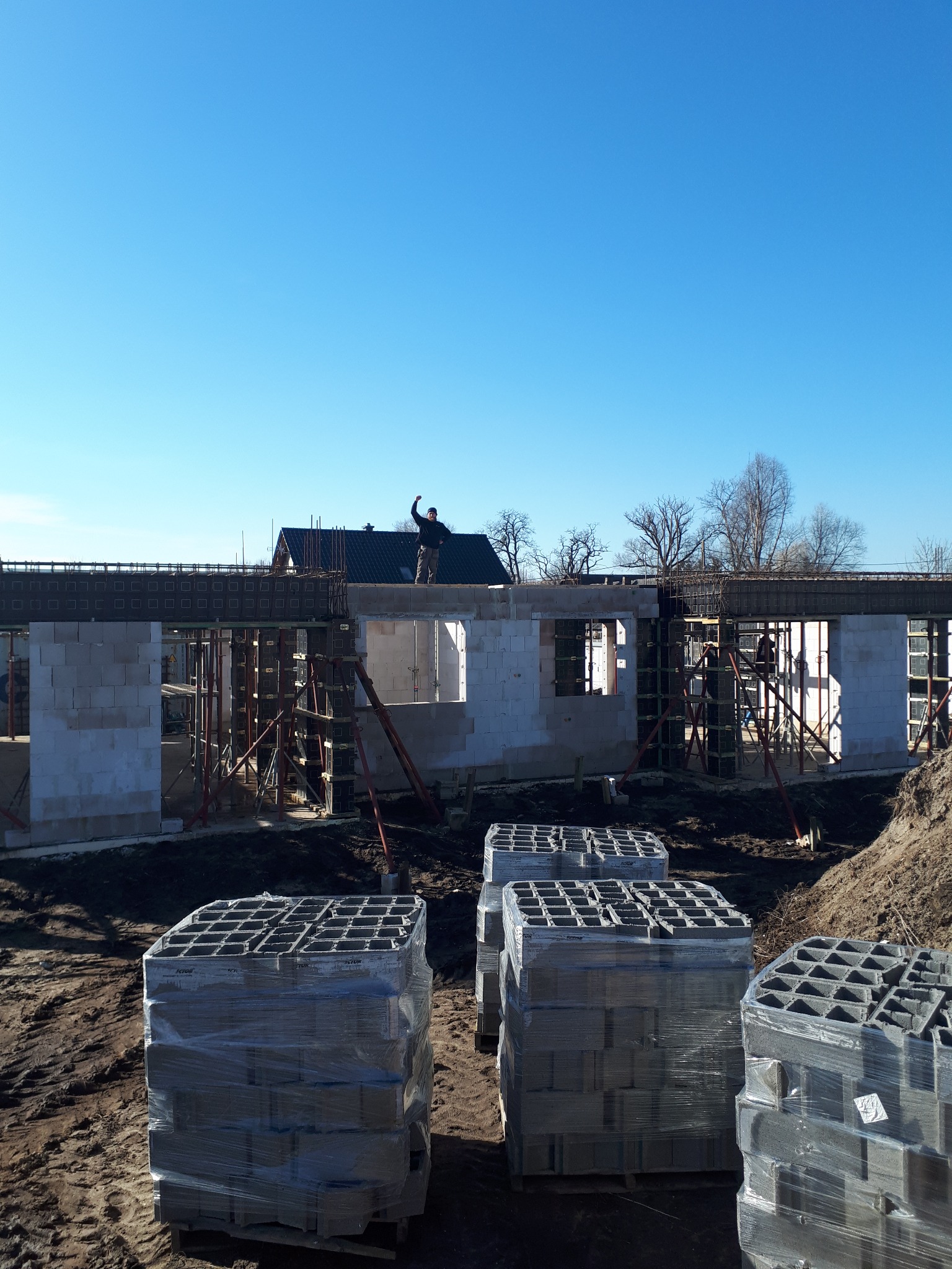 Wznoszenie ścian parteru domu z betonu komórkowego Ytong, widoczne zbrojenie nadproży, palety bloczków na pierwszym planie, pracownik na górze budynku macha ręką.