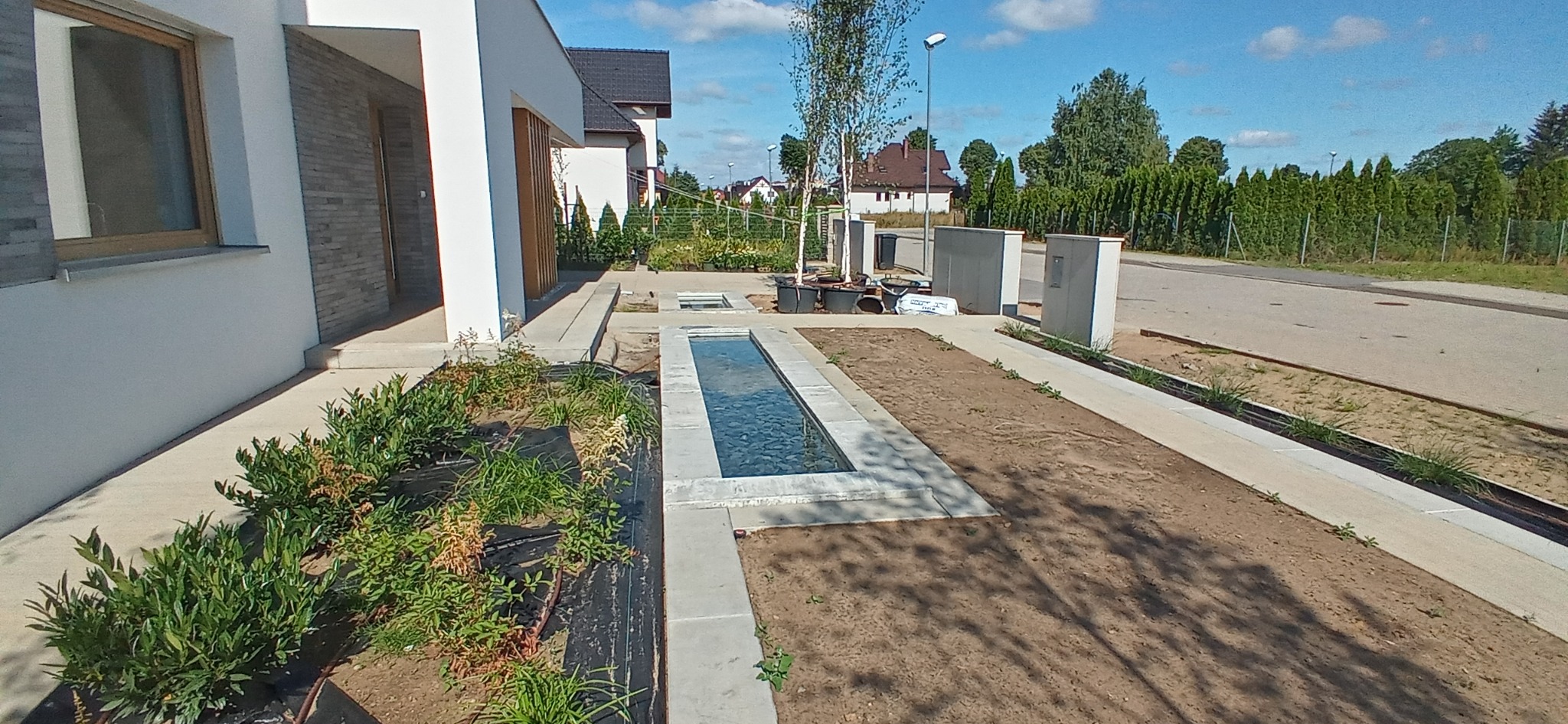 Nowoczesny dom jednorodzinny z elementami zagospodarowania terenu: podłużny zbiornik wodny z kamieniami, świeżo nasadzone rośliny i trawnik w trakcie zakładania.