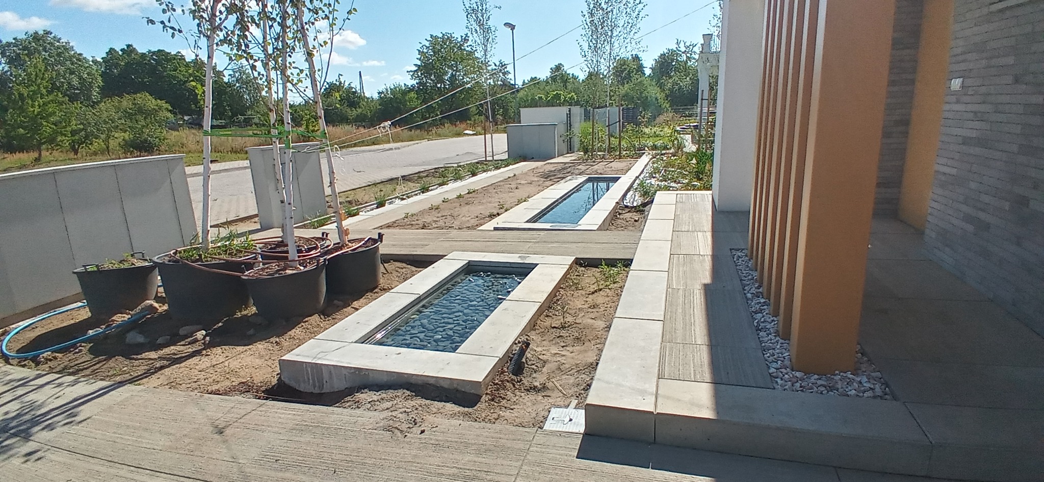 Nowoczesny ogród z minimalistycznymi, prostokątnymi zbiornikami wodnymi wypełnionymi kamieniami, świeżo posadzone drzewa w donicach, jasna kostka brukowa, elementy drewniane i kamienne, w tle dom...