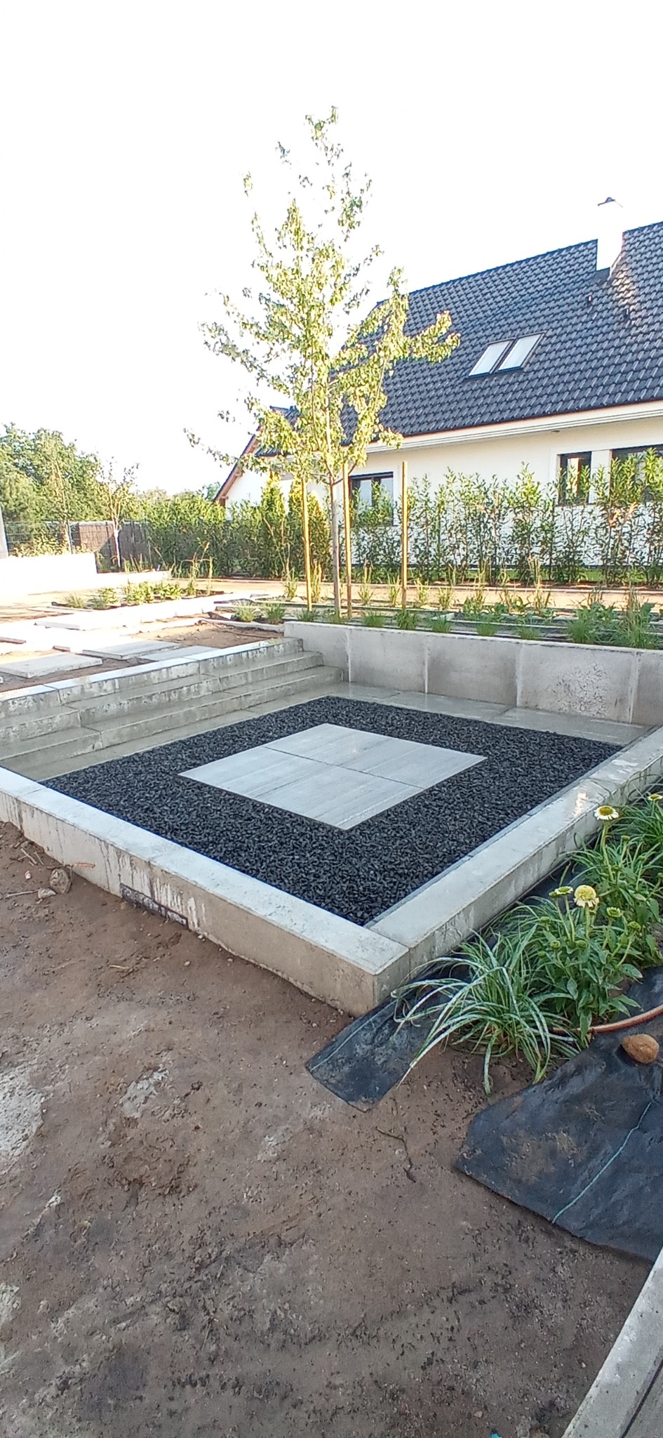 Nowoczesny taras z betonową ramą, wypełniony czarnym żwirem i kwadratowymi płytami, w tle dom jednorodzinny z czarnym dachem.