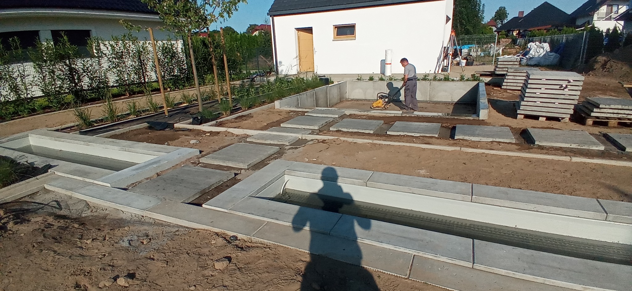 Prace ziemne i betonowe przy budowie ogrodu z elementami wodnymi; pracownik zagęszcza podłoże ubijarką wibracyjną, widoczne betonowe koryta i płyty chodnikowe.