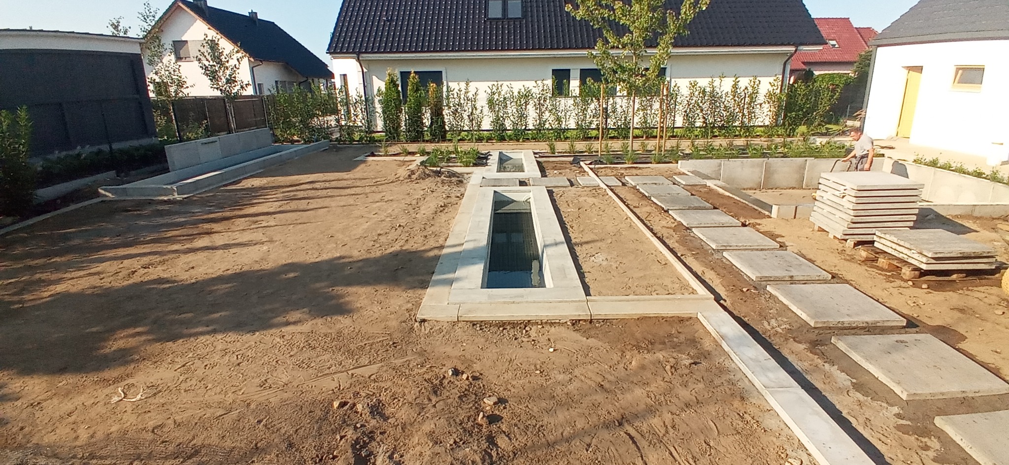 Wykopane, betonowe zbiorniki na wodę deszczową w ogrodzie, częściowo zakryte płytami betonowymi, w tle domy jednorodzinne i zieleń.