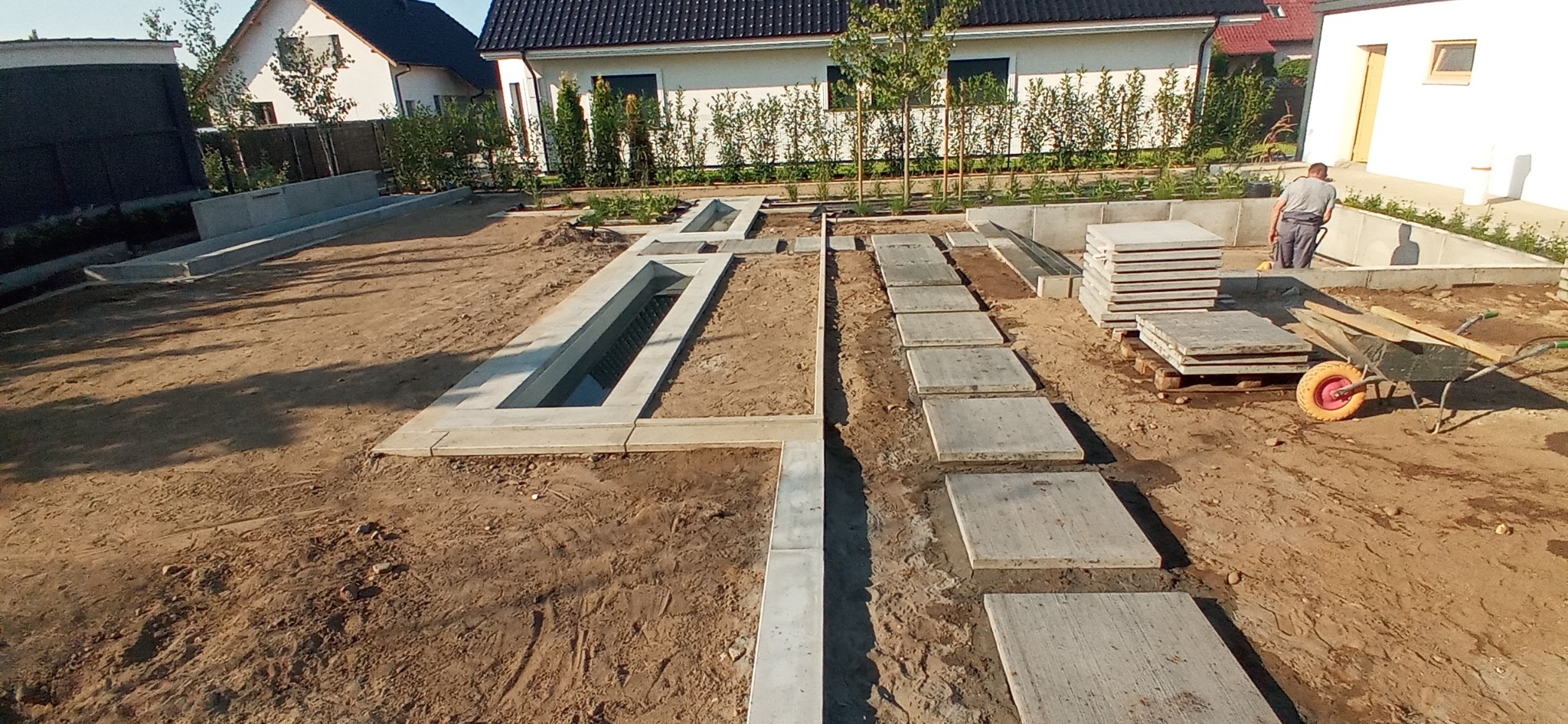 Wykop pod fundamenty domu jednorodzinnego z częściowo wylanymi betonowymi ścianami, widoczne płyty chodnikowe ułożone jako tymczasowa ścieżka i pracownik z taczką w tle.