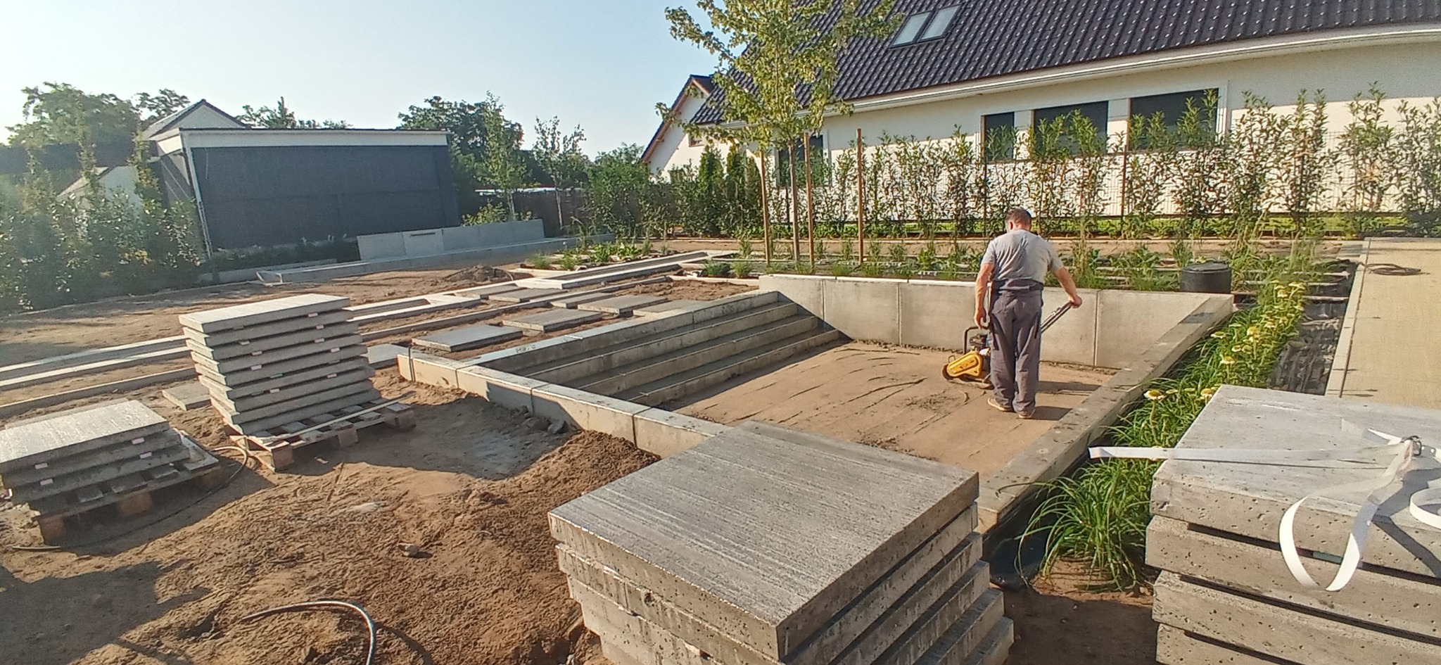 Prace ziemne przy budowie stawu: ubijanie podłoża w wykopie z betonowymi obrzeżami, stosy płyt betonowych obok, w tle budynki mieszkalne i zieleń.