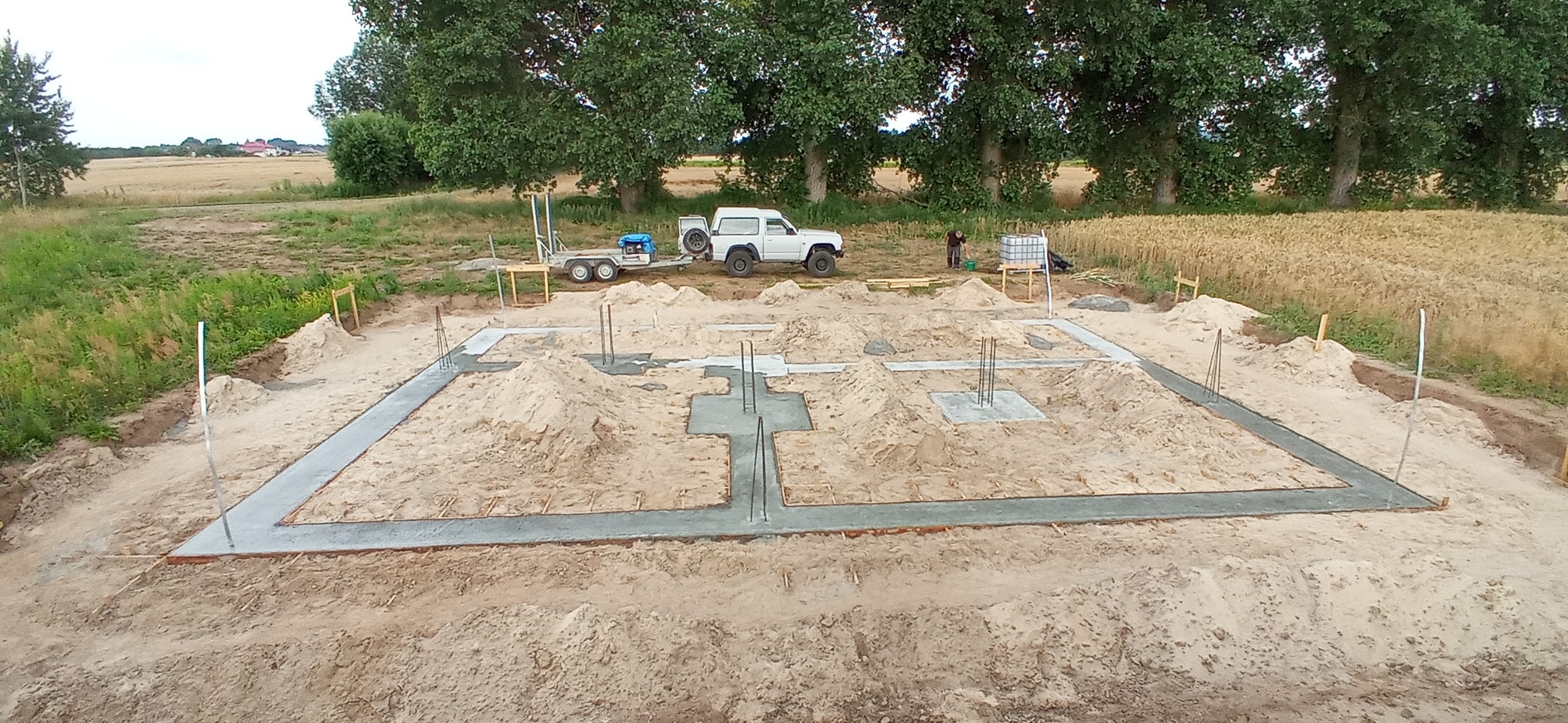Wylane fundamenty domu jednorodzinnego z wystającymi prętami zbrojeniowymi, na działce z polami uprawnymi w tle, widoczny samochód terenowy z przyczepką i pracownik budowlany.