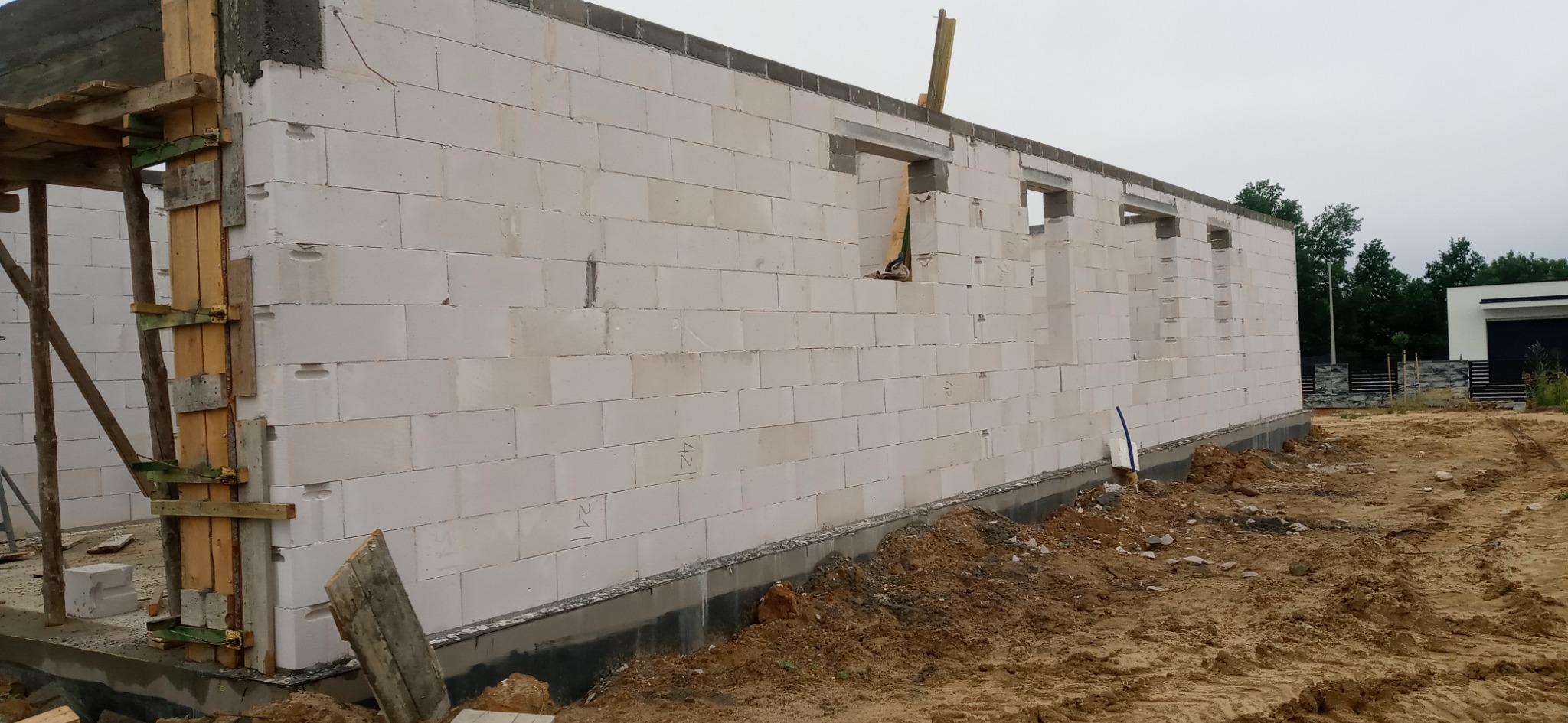Wznoszenie ścian parteru domu z białych bloczków betonowych, widoczne otwory okienne, szalunki narożne i fundament zabezpieczony izolacją.
