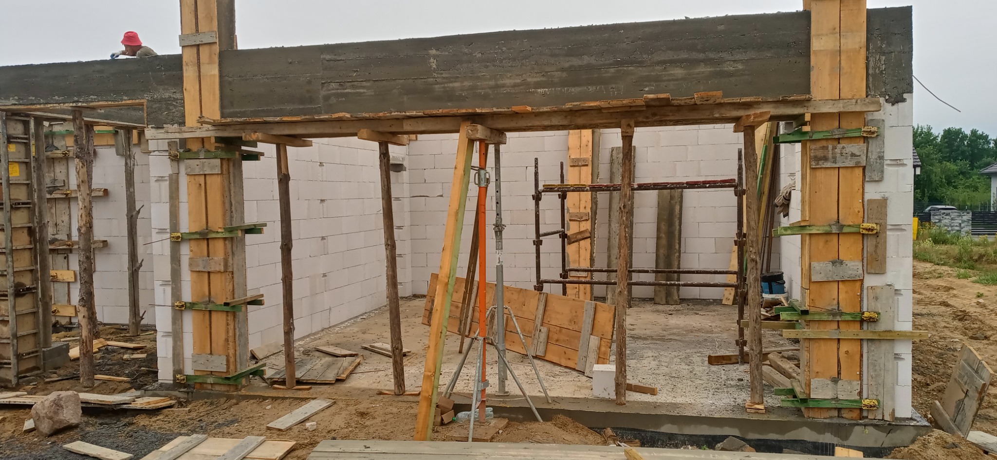 Wznoszenie ścian domu z betonu komórkowego, widoczne szalunki drewniane i podpory, w tle pracownik w czerwonym kapeluszu na górnej belce.