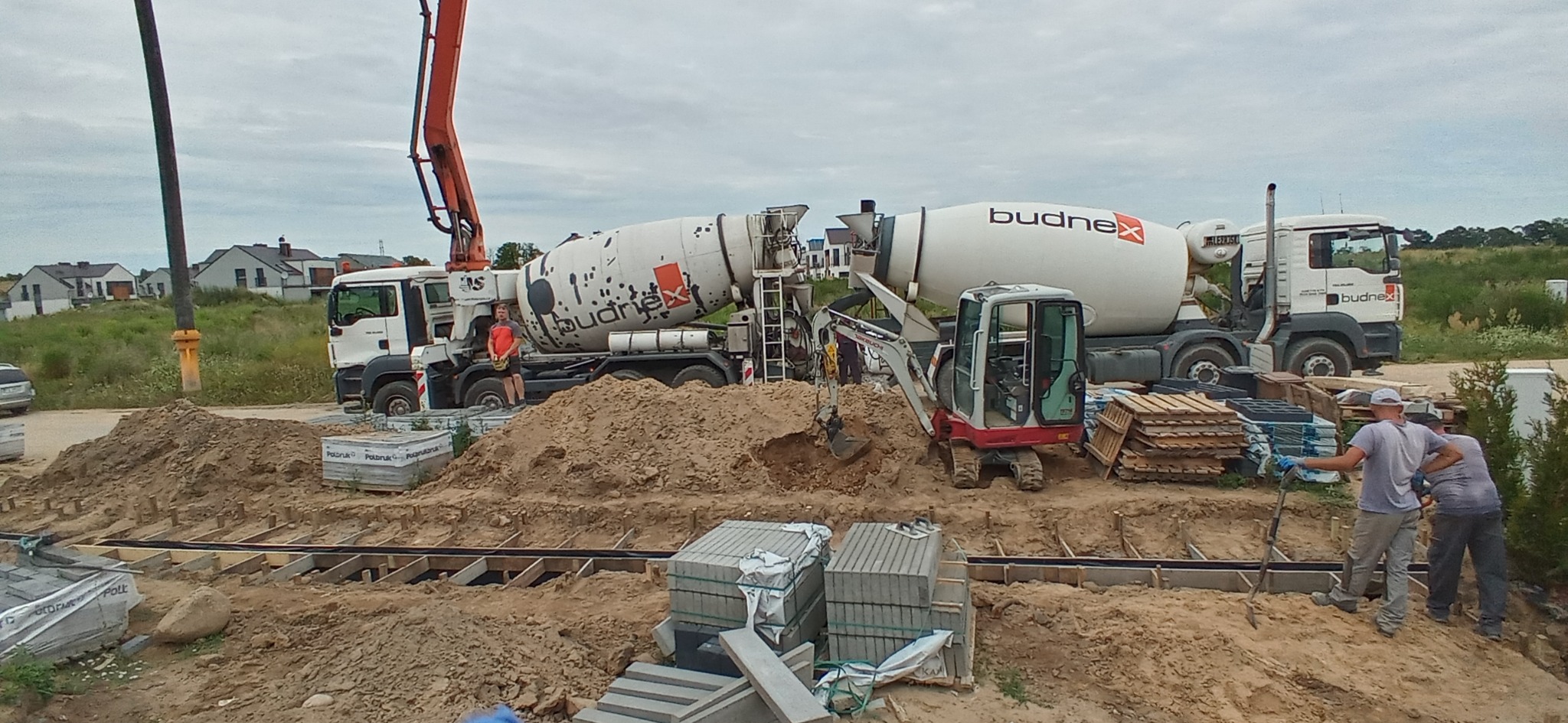 Dwa betonomieszarki firmy Budnex na placu budowy fundamentów, pracownicy przy wykopie, w tle mini koparka i nowe domy.