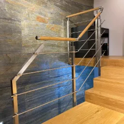 Schody z drewnianymi stopniami i balustradą ze stali nierdzewnej z drewnianymi elementami, na tle ściany wykończonej płytami z naturalnego kamienia.