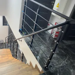 Widok z góry na schody z drewnianymi stopniami i czarną balustradą z poziomymi prętami, marmurowa podłoga, biała ściana, drzwi w tle, skrzynka elektryczna z ostrzeżeniem.