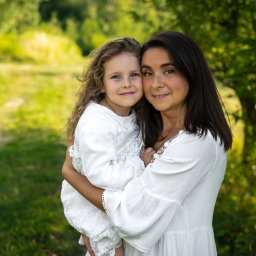 Oczami Zajawki Fotografia - Portret matki z córką w białych sukienkach na tle zieleni. Uśmiechnięte twarze, naturalne światło, miękkie tło. Romantyczna atmosfera, bliskość i ciepło rodzinne.